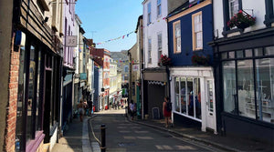 A photo of the falmouth high street with rows of colourful shops either side and a narrow road in between. Bunting hangs between them and people are walking past.